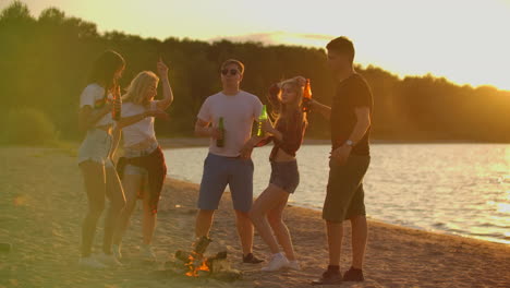 The-young-students-spend-summer-evening-at-sunset-on-the-beach-in-shorts-and-t-shirts-around-bonfire-with-beer.-They-are-dancing-and-enjoying-the-view-in-nature.